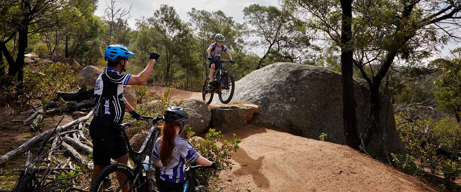 A young mountain biker attempts a drop while cheered on by his father and older sister.