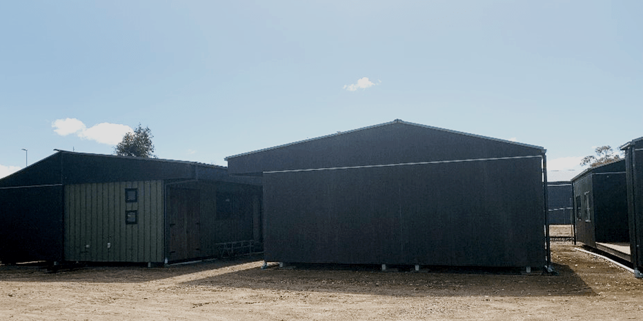 Back and front view of two cabins with a third cabin showing a built-in outdoor seating area. The cabins have black roofs and green coloured timber cladding. There are black framed windows in the walls.