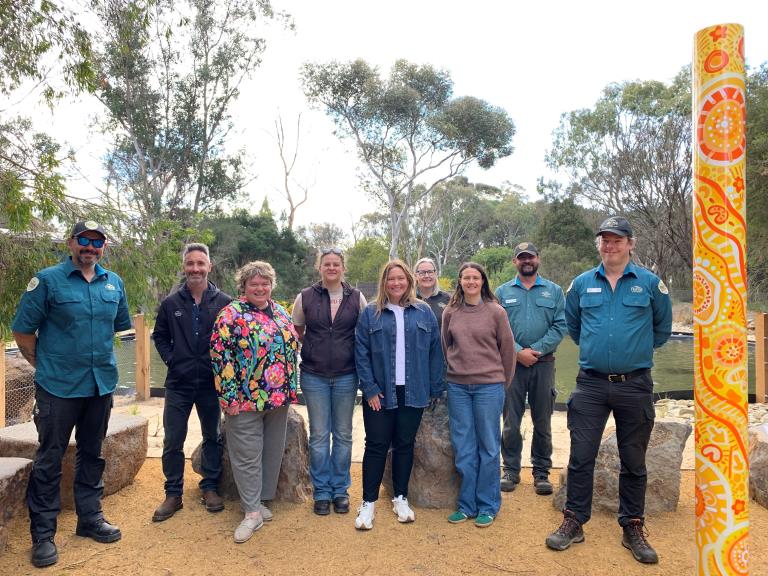 Smiling group of people standing on ramped earth in front of the Serendip display pond. A tall pole at right features a yellow, orange and white artwork by Wadawurrung artist Jenna Oldaker. This was taken when the Stakeholder Reference Group visited the almost completed Sensory Garden.