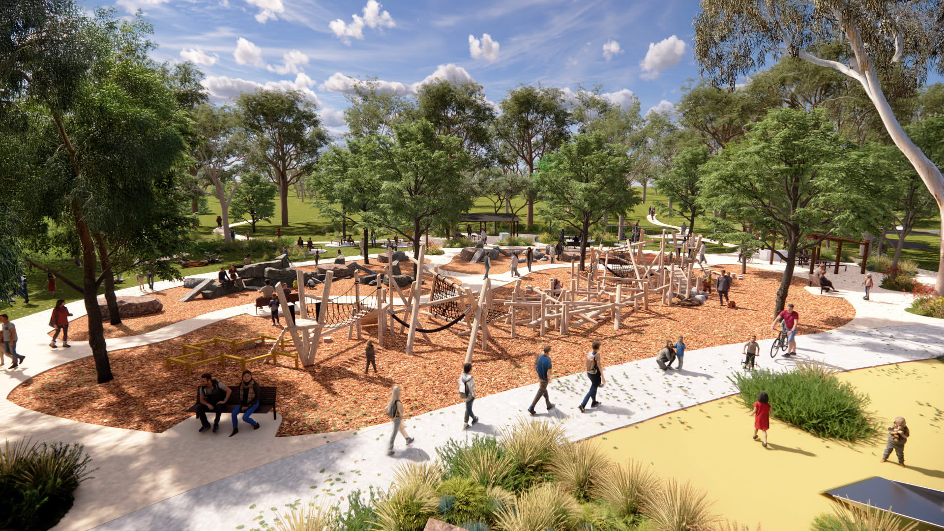 Open nature themed play area with walking path circling play equipment and tall native trees behind. People are walking on the park and relaxing in the sun under trees. Children are also playing on the play equipment as the sun shines and clouds dot the blue sky.