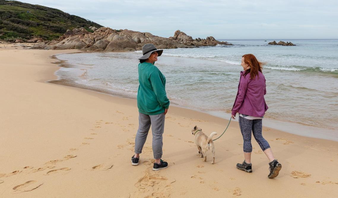 Two women walk at the waters edge along West Cape Beach at Cape Conran Costal Park with Salmon Rocks in the Background.