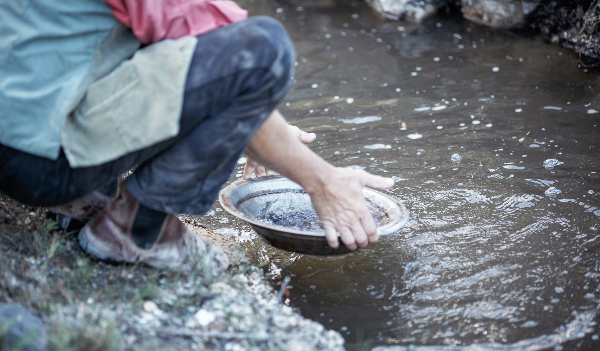 A boy crouches at the waters panning for gold in a river.