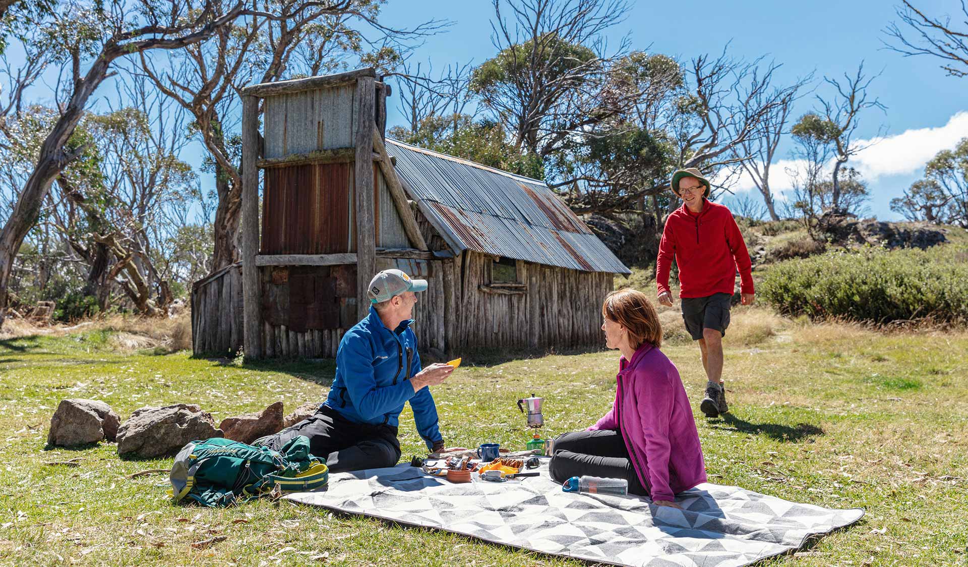 Three friends enjoy a picnic on the grass in front of the historic Wallace Hut.
