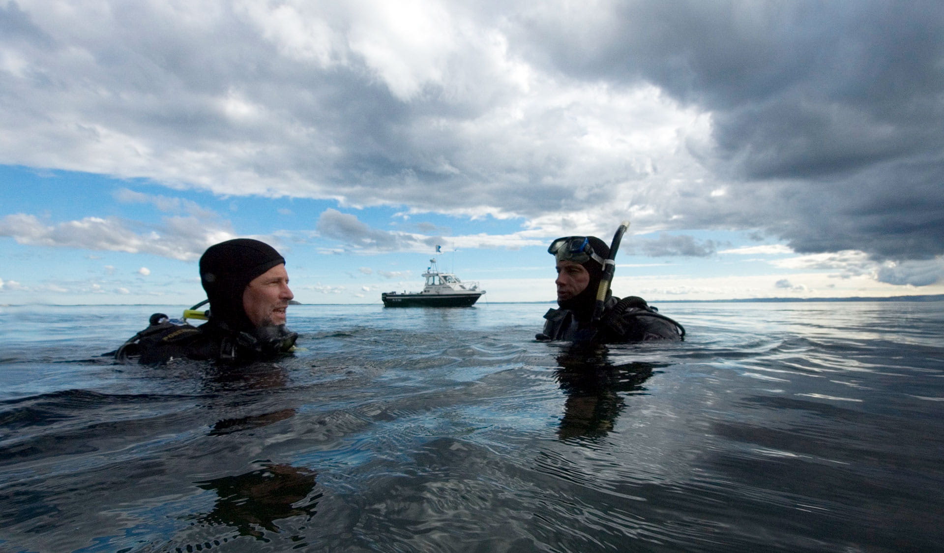 Two male divers return to the water's surface during their dive with their boat in the background of the shot.