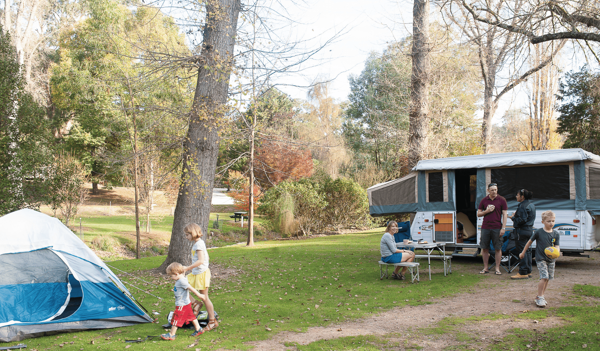 Two kids walking towards tent, while adults talk with ranger near caravan at campground.