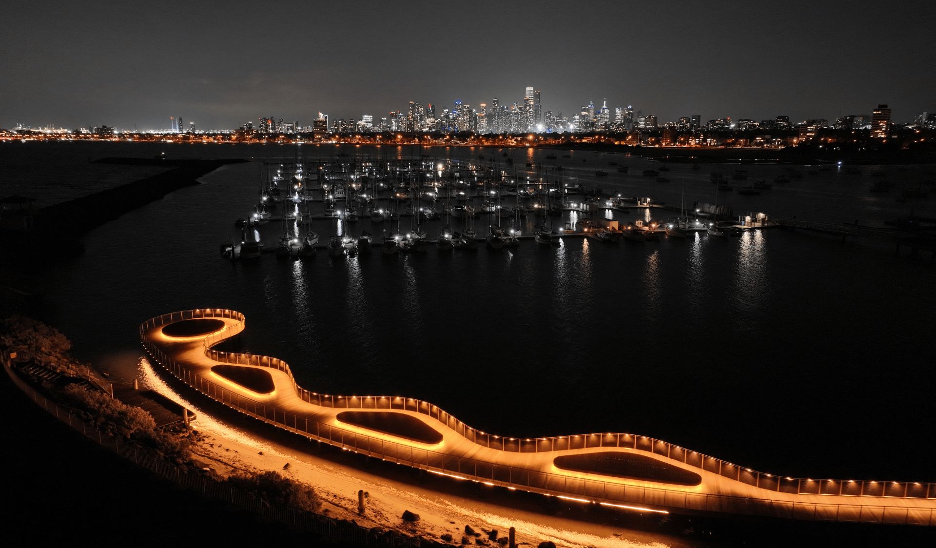 View of St Kilda Pier lit up at nighttime with city skyline in the background.