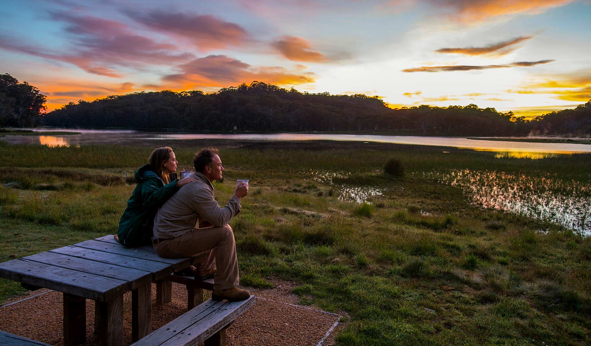 A couple watch the sunset at a picnic table on the edge of Lake Catani at Mount Buffalo National Park.