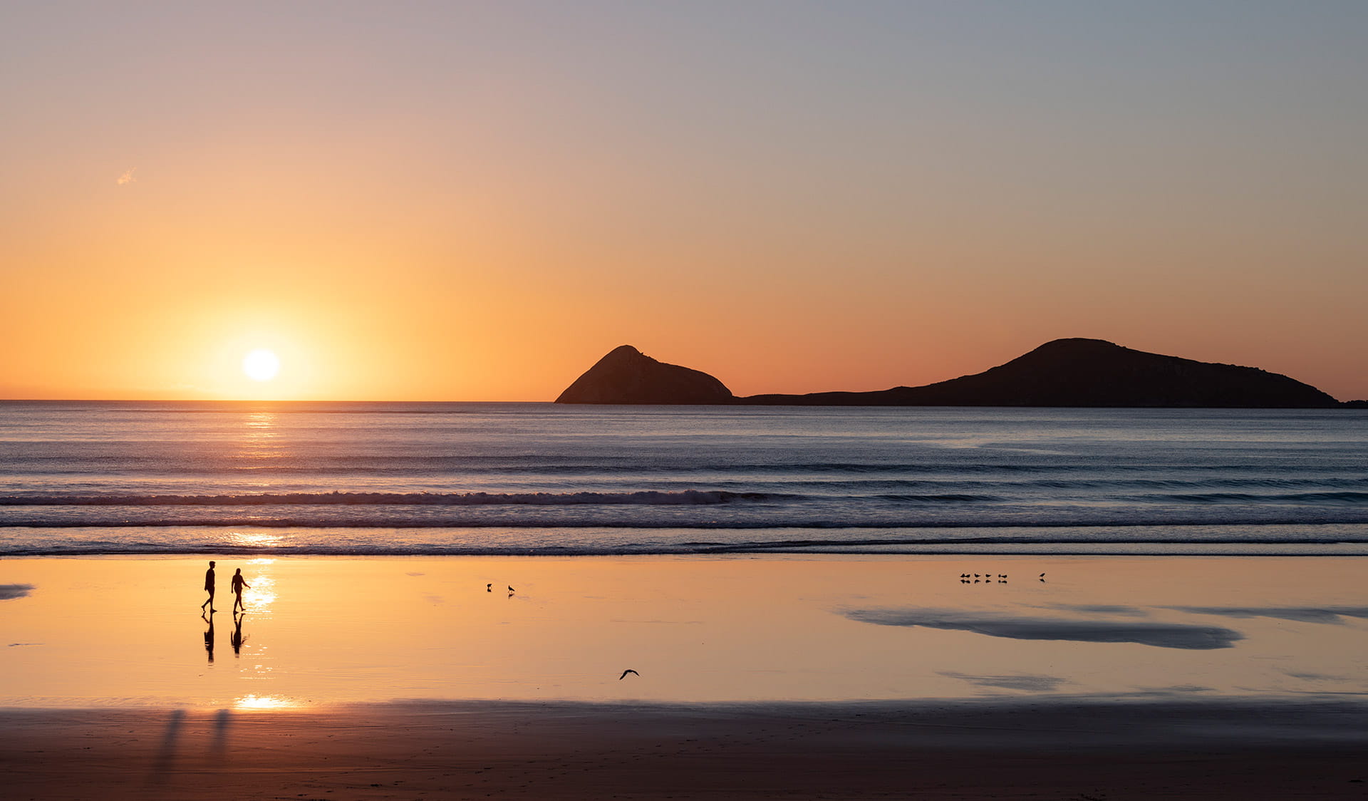 Two walkers walk along a beach at Wilsons Promontory as the sun dips below the horizon. 