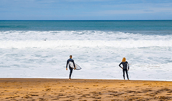 Two men in wetsuits hold surfboards as they look out over the ocean