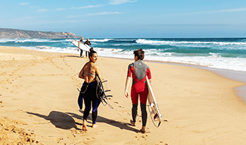 A woman and a boy walk towards the surf while holding surfboards. The woman looks back at the camera