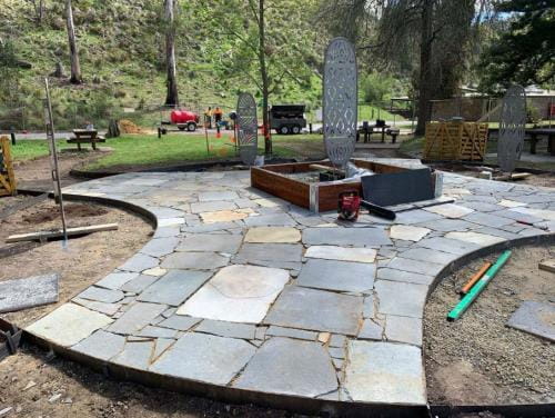 Workers stand behind blue and sand colour pavers which have just been laid in the Yarning Circle. There are Aboriginal symbols etched on three metal panels one in the middle and tow behind. There is a green grass hill and trees in the background